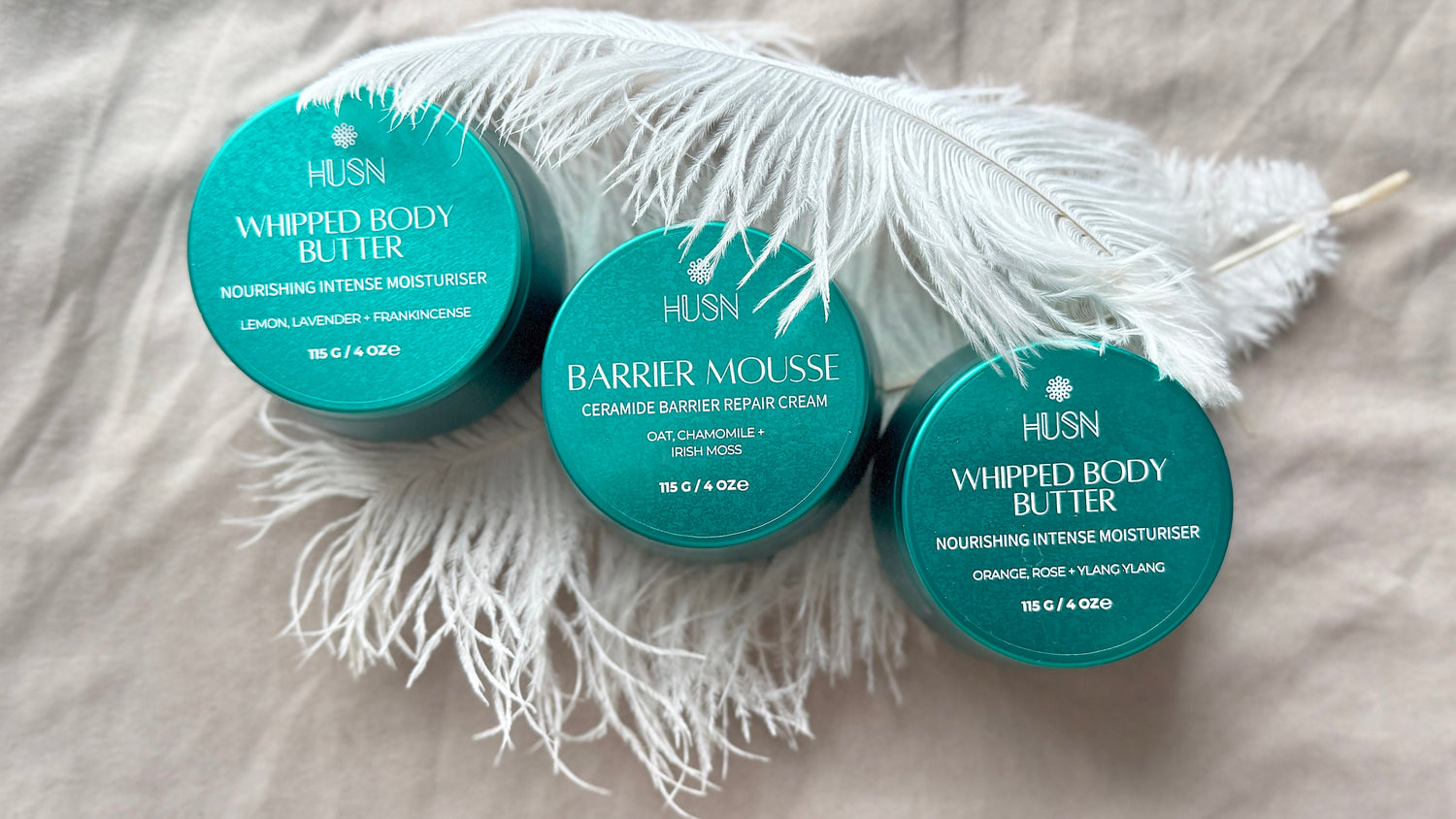 Three containers of HUSN body butter on a textured surface with feathers.